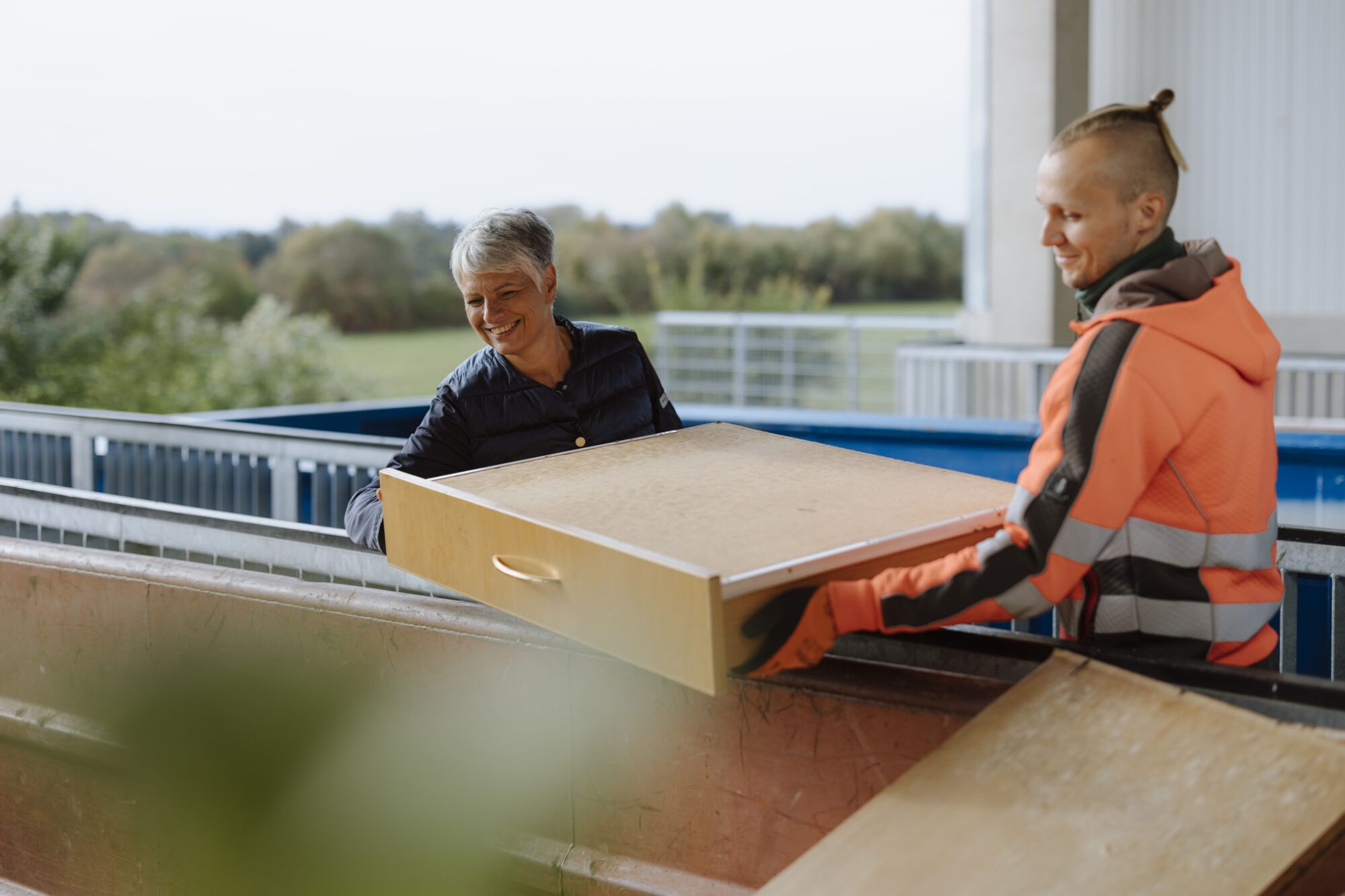 Eine ältere Dame mit grauem Haar und einer blauen Jacke, sowie ein Mitarbeiter des Sammelzentrum, gekleidet in eine orange Warnwestenjacke sowie orange Handschuhe, halten gemeinsam eine Lade eines Schranks über einen Sperrmüllcontainer.