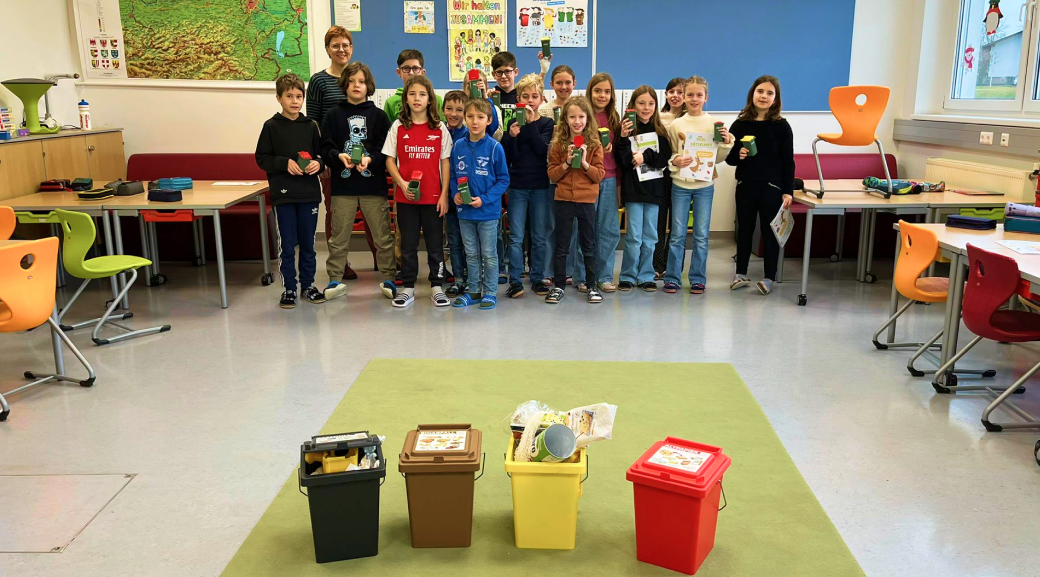 Schulklasse mit kleinen Mülltonnen in den Händen im Hintergrund; im Vordergrund Tonnen für Restmüll, Gelbe Tonne, Altpapier- und Biotonne