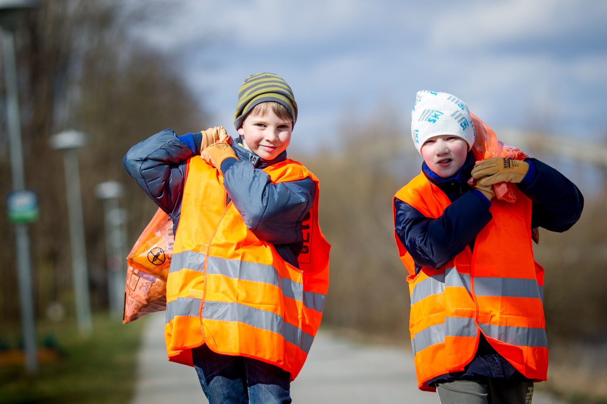 Zwei Kinder gehen mit Warnwesten und Müllsäcken auf dem Rücken