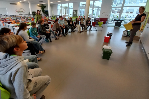 Schulkinder sitzen in einem Sessel-Halbkreis und die Abfallberaterin aus dem Bezirk Scheibbs hält eine kleine Gelbe Tonne in der Hand und spricht zu den Kindern.