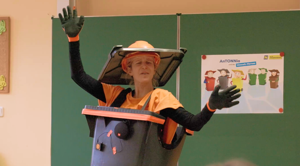 Zu sehen ist eine als Restm&uuml;lltonne verkleidete Schauspielerin, die in einer Schulklasse vor der Tafel singt.