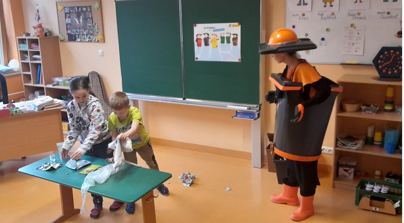2 Kinder legen die Abfälle, die am Boden liegen, auf einen Tisch, AnTONNia steht daneben