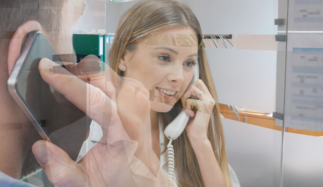Mann hält Handy ans Ohr. Frau hält Telefonhörer ans Ohr. Im Hintergrund ein Büro.