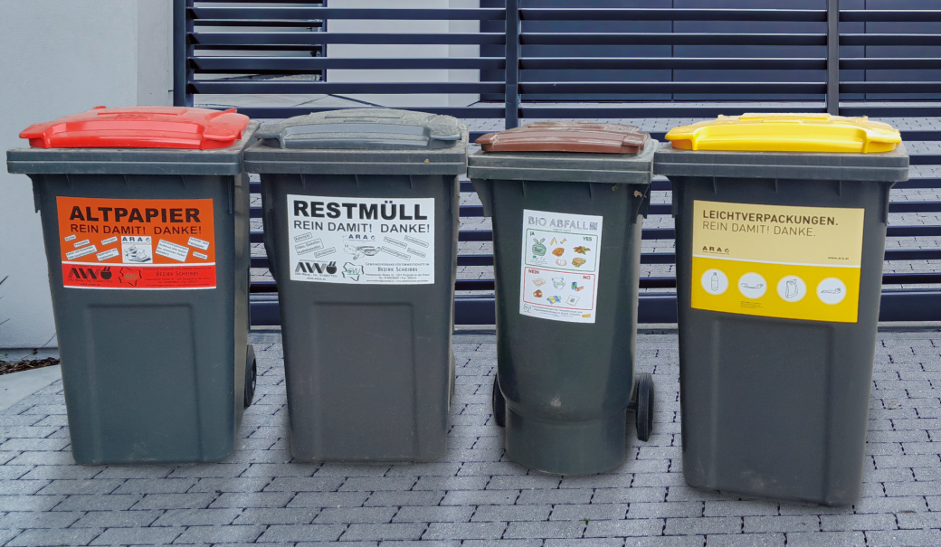 4 Abfalltonnen (roter Deckel für Altpapier, schwarzer Deckel für Restmüll, brauner Deckel für Bioabfälle und gelber Deckel für Verpackungen) stehen vor einer Hauseinfahrt