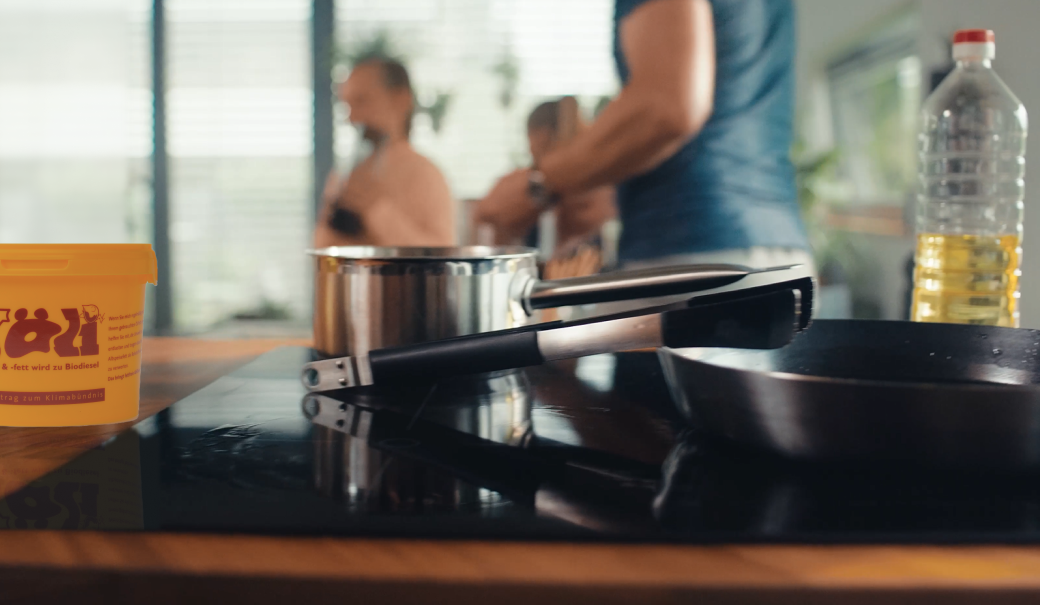K&uuml;chenherd mit Bratpfanne und Speise&ouml;lflasche rechts und N&Ouml;LI links im Bild; im Hintergrund unscharf: Glasscheiben und Personen